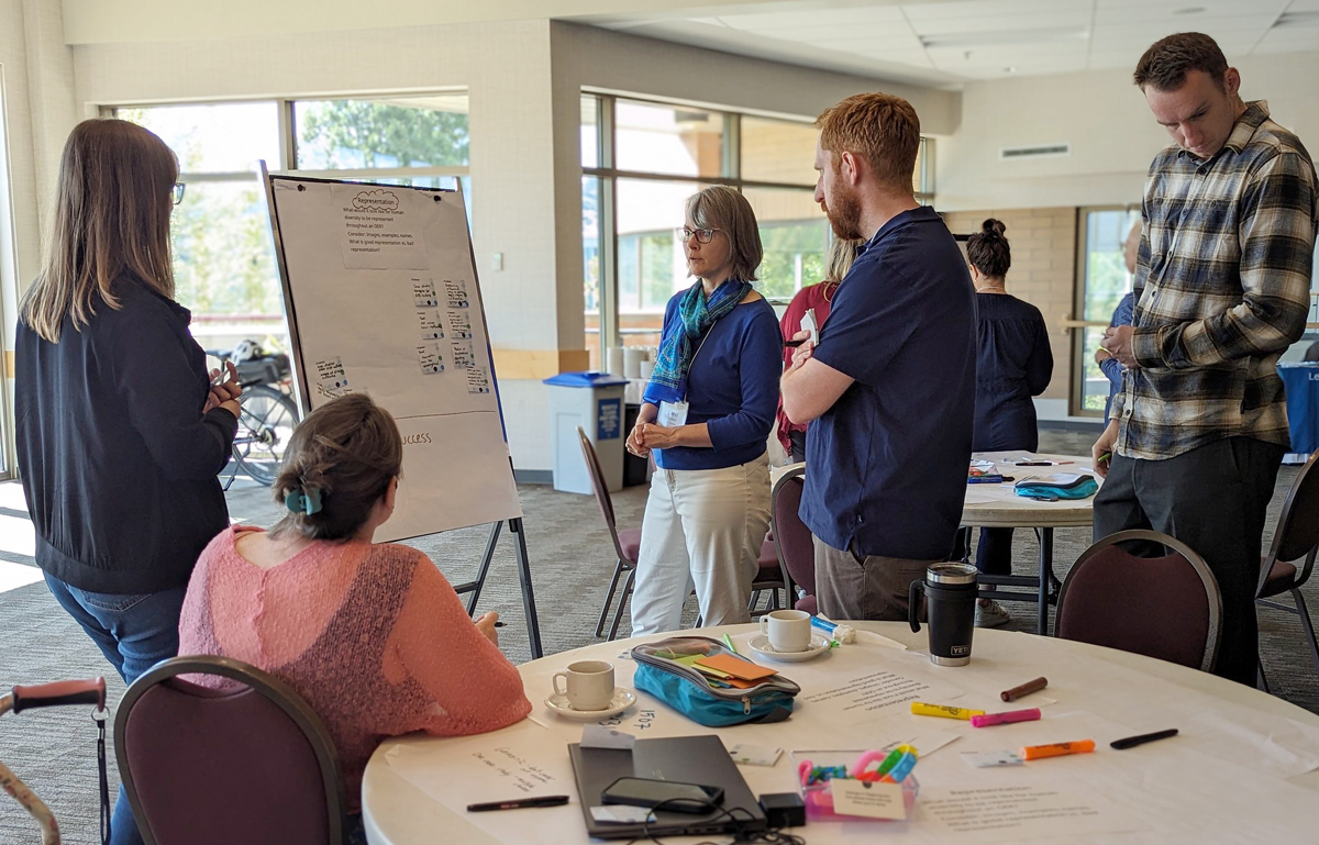 Five people gather around an easel. They are in conversation and appear to be brainstorming. They are in a meeting room with a round table and large windows in the background.