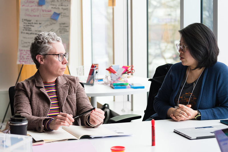 Two people look directly at each other in conversation. They are sitting at a desk in a brightly lit room with windows. One has short grey hair and a brown jacket. The other has shoulder-length dark hair and is wearing a dark blue sweater.