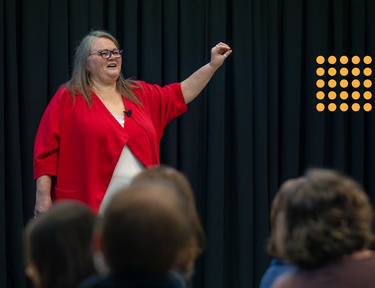 Jo Chrona presents to the audience in front of a black curtain. She is wearing a red top and is smiling with her arm raised.