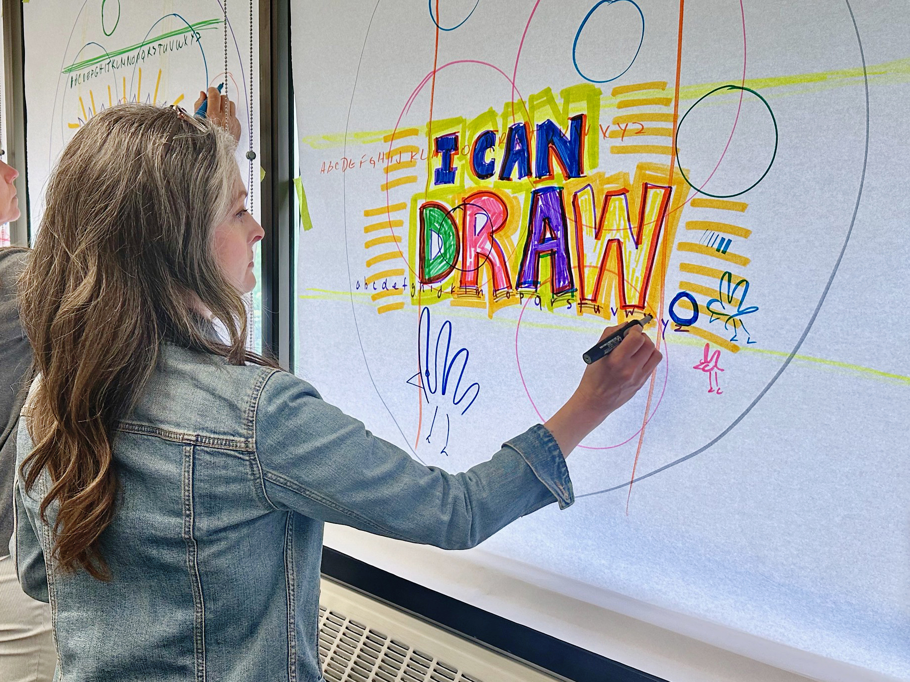 A person with long hair wearing a denim jacket draws on poster paper. The poster reads 'I CAN DRAW'.